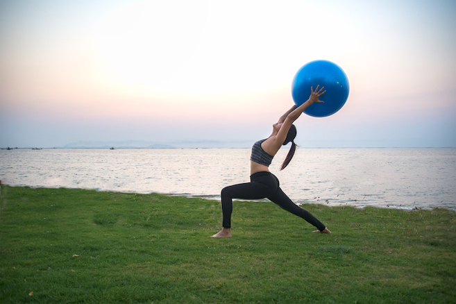 woman doing a partial backbend while holding a workout ball on a grassy field