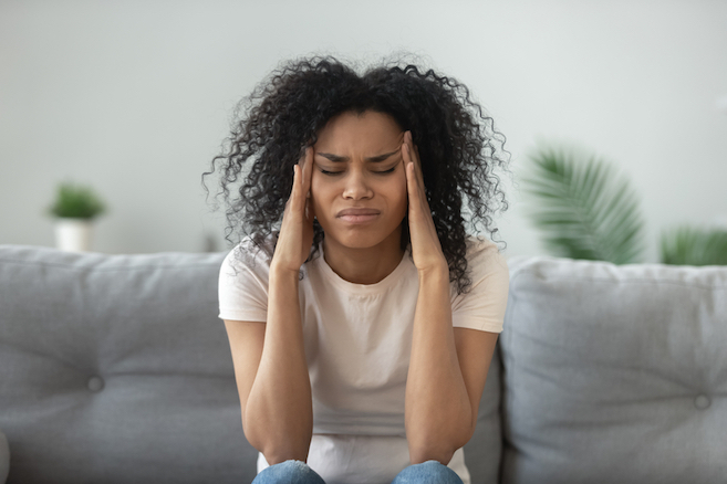 black woman sitting on the couch holding her head with a migraine