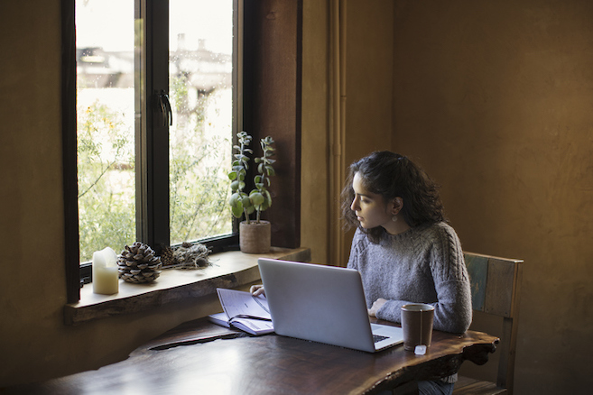 Sitting next to a window, embraced by natural light, this determined young adult (mixed-race) woman is working from home on a tight deadline. She is leaning in and focused to finish her project. Prominent laptop computer, a reference book, and mug of tea on a natural-edged redwood slab table complete her workspace. A houseplant, pine cone, and candle sit on the window sill. Outdoors: yellow flowers, foliage and daylight are visible in soft focus. Young woman wears a cozy gray wool sweater and has dark brown, wavy hair. Natural light illuminates her workspace and her profile. She appears to be turning the page of the reference book, while reading.