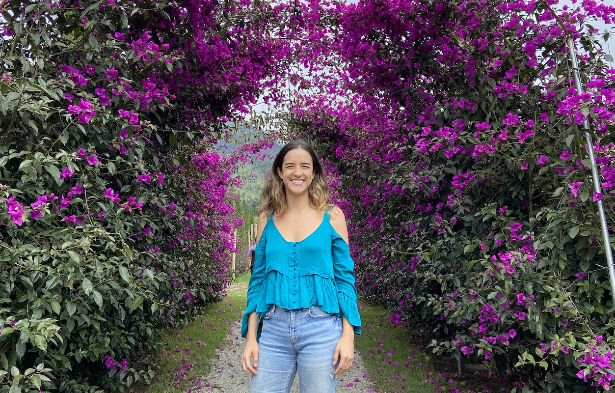 Photo of Amalia Arango in a blue shirt standing in front of trees with pink flowers